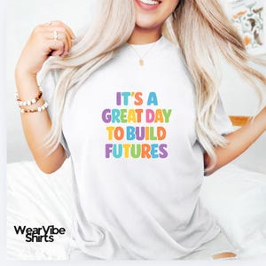 May include: White t-shirt with the text "IT'S A GREAT DAY TO BUILD FUTURES" in colorful letters. The shirt has a crew neck and short sleeves. The WearVibe Shirts logo is at the bottom left.