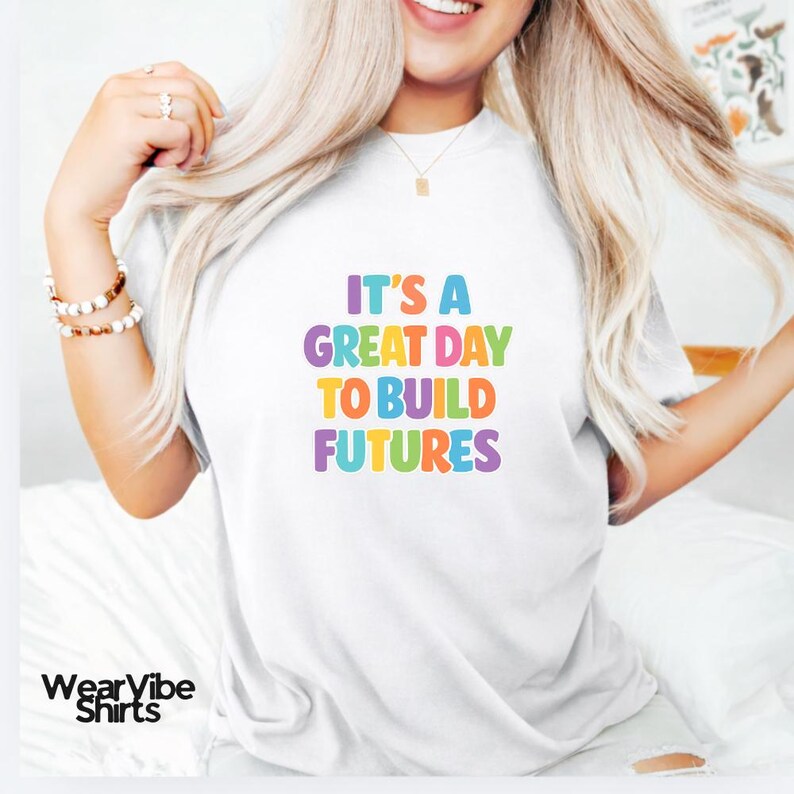 May include: White t-shirt with the text "IT'S A GREAT DAY TO BUILD FUTURES" in colorful letters. The shirt has a crew neck and short sleeves. The WearVibe Shirts logo is at the bottom left.