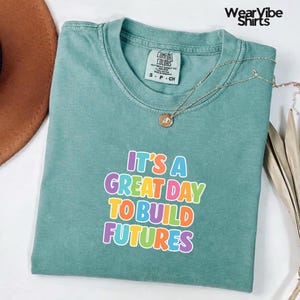 May include: A teal Comfort Colors t-shirt with the text "IT'S A GREAT DAY TO BUILD FUTURES" in colorful letters. A silver necklace with a small pendant is draped across the shirt. The shirt is a size small.