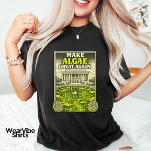 May include: A black t-shirt featuring a graphic print of the Lincoln Memorial surrounded by a green algae-filled pond. The text reads "MAKE ALGAE GREAT AGAIN" in yellow and white letters. A small white duck swims in the centre of the pond.