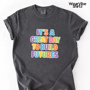 May include: Dark gray t-shirt with rolled-up sleeves. The shirt features the text "IT'S A GREAT DAY TO BUILD FUTURES" in colorful bubble letters. The brand name "WearVibe Shirts" is printed in the upper right corner.