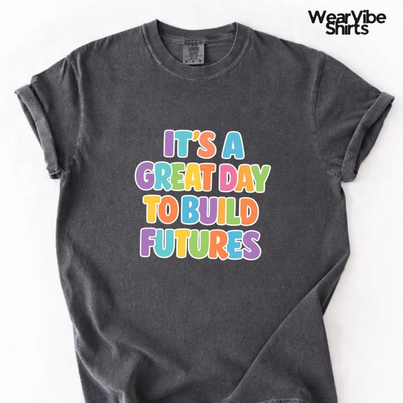 May include: Dark gray t-shirt with rolled-up sleeves. The shirt features the text "IT'S A GREAT DAY TO BUILD FUTURES" in colorful bubble letters. The brand name "WearVibe Shirts" is printed in the upper right corner.