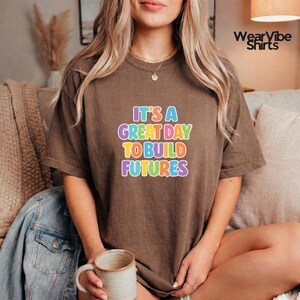 May include: A brown t-shirt with the text "IT'S A GREAT DAY TO BUILD FUTURES" in colorful, rainbow-colored letters. The shirt is a neutral color and has a relaxed fit. The shirt is a casual wear item.