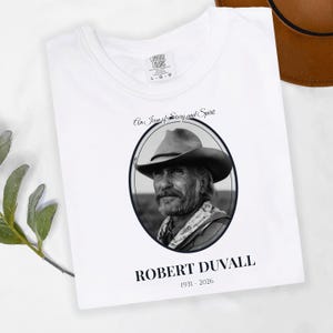 May include: White t-shirt featuring a black and white portrait of Robert Duvall in an oval frame. The text "An Icon of Story and Spirit" and "ROBERT DUVALL 1931-2026" are printed on the shirt. The shirt is a Comfort Colors L-G-G.