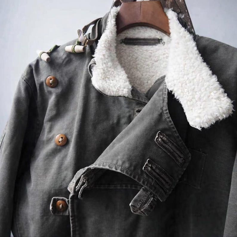 May include: A dark gray jacket featuring a white faux shearling collar and lining. The jacket includes brown circular buttons, toggle-style closures with green cord accents, and a double-breasted front design with a structured collar and lapels.