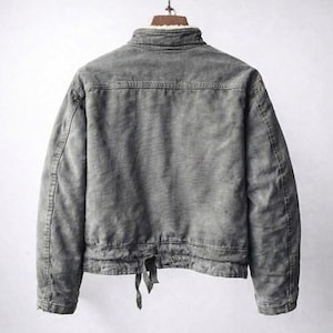 May include: A charcoal gray jacket with a weathered texture, featuring a high collar lined with white faux fur. The back displays a horizontal yoke seam, long sleeves, and a bottom hem with an adjustable tie closure hanging from the center.