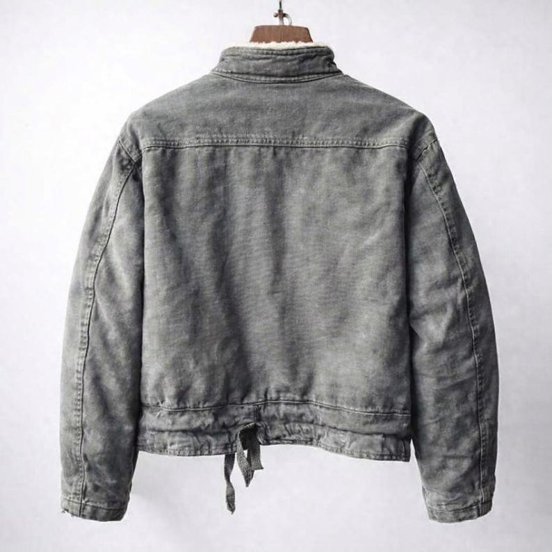 May include: A charcoal gray jacket with a weathered texture, featuring a high collar lined with white faux fur. The back displays a horizontal yoke seam, long sleeves, and a bottom hem with an adjustable tie closure hanging from the center.