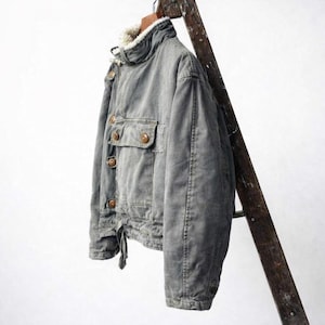 May include: A grey jacket featuring a high collar lined with white faux fur, a front button closure with brown buttons, and a single chest pocket with a buttoned flap. The jacket has long sleeves and a cinched hemline with a visible fabric tie.