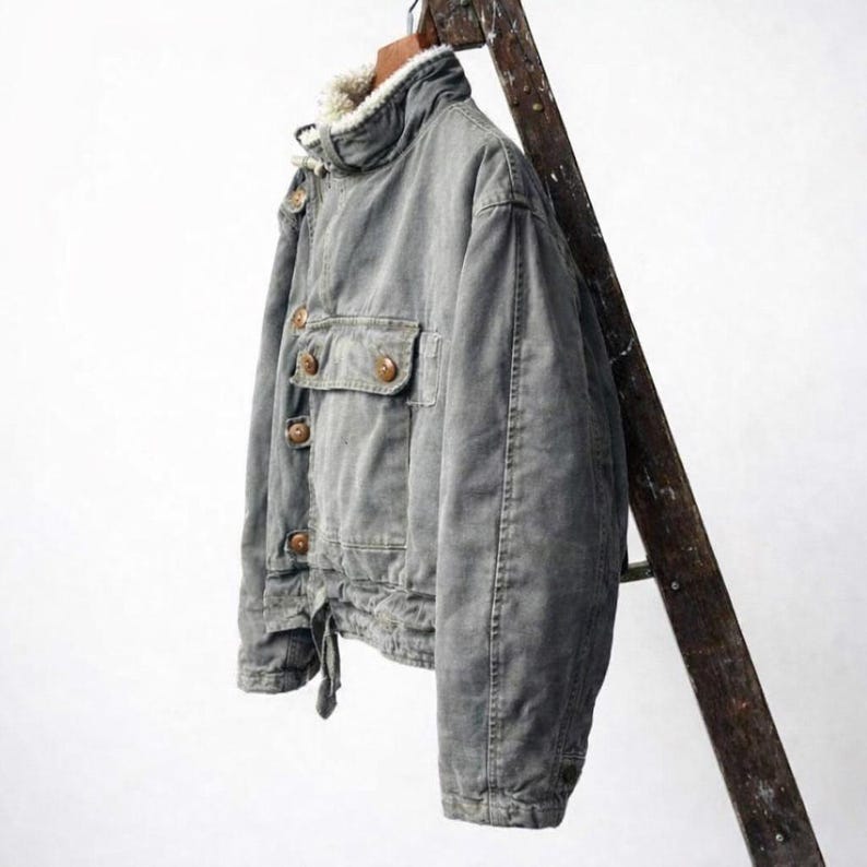 May include: A grey jacket featuring a high collar lined with white faux fur, a front button closure with brown buttons, and a single chest pocket with a buttoned flap. The jacket has long sleeves and a cinched hemline with a visible fabric tie.