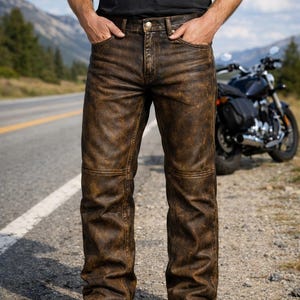 May include: Distressed brown leather pants featuring a straight-leg cut, reinforced horizontal knee stitching, a standard button fly closure, and five-pocket styling. The finish displays a weathered, textured appearance with darker brown shading.