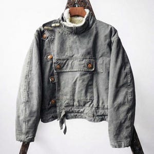 May include: A grey canvas jacket featuring a high collar lined with white faux shearling, an asymmetrical front closure with wooden toggle buttons, and a large rectangular chest pocket. The jacket has long sleeves and a straight hemline.