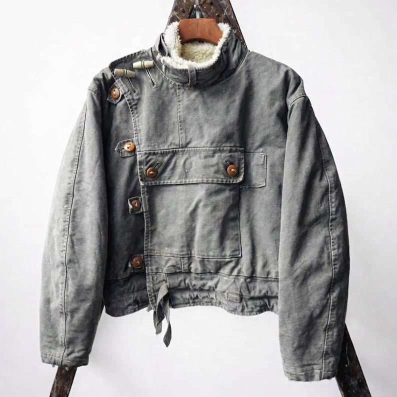 May include: A grey canvas jacket featuring a high collar lined with white faux shearling, an asymmetrical front closure with wooden toggle buttons, and a large rectangular chest pocket. The jacket has long sleeves and a straight hemline.