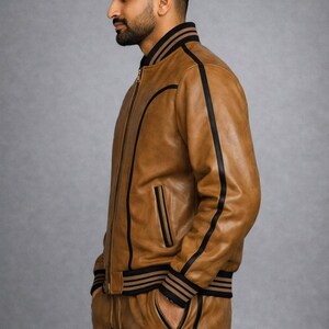 Pu&ograve; includere: Giacca bomber e pantaloni coordinati in pelle marrone chiaro, con strisce e finiture nere. La giacca ha una chiusura con cerniera e tasche. I pantaloni hanno una vita con coulisse e tasche con cerniera. L'outfit &egrave; mostrato su sfondo grigio.