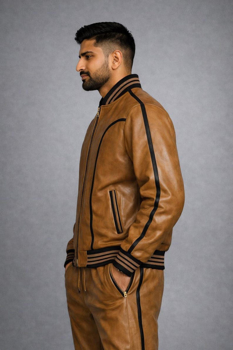 Pu&ograve; includere: Giacca bomber e pantaloni coordinati in pelle marrone chiaro, con strisce e finiture nere. La giacca ha una chiusura con cerniera e tasche. I pantaloni hanno una vita con coulisse e tasche con cerniera. L'outfit &egrave; mostrato su sfondo grigio.