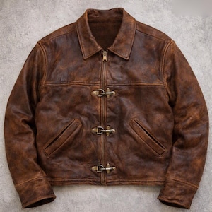 May include: A brown distressed leather jacket featuring a classic collar, long sleeves, and a central zipper closure. The front is accented with three horizontal metal toggle clasps positioned over the zipper, along with two slanted side pockets.