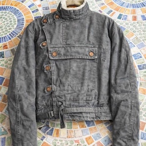 May include: A charcoal gray jacket with a high collar lined in cream-colored faux fur. The jacket features an asymmetrical front closure with brown buttons, a large rectangular chest pocket with two buttons, and a waist strap with a buckle.