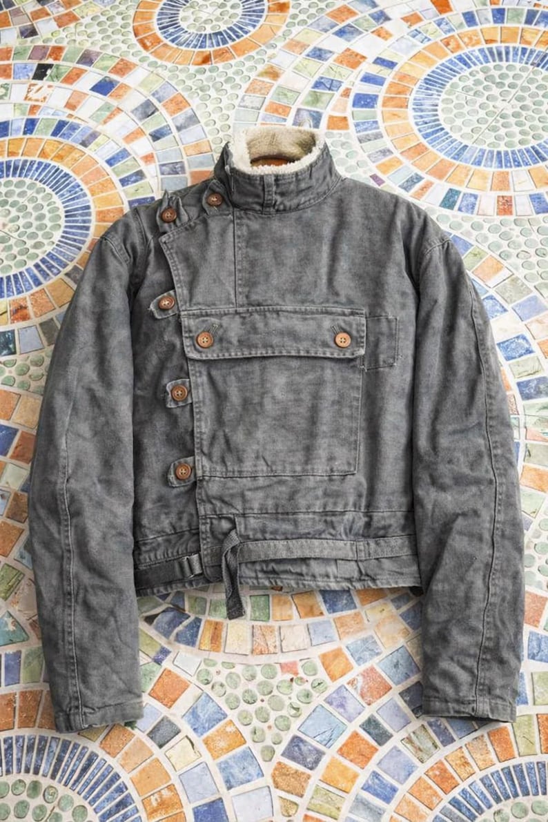 May include: A charcoal gray jacket with a high collar lined in cream-colored faux fur. The jacket features an asymmetrical front closure with brown buttons, a large rectangular chest pocket with two buttons, and a waist strap with a buckle.