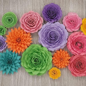 May include: A collection of colorful paper flowers in various sizes and styles. The flowers are in shades of pink, purple, orange, green, yellow, and teal. The flowers are arranged on a light wood-grain surface.