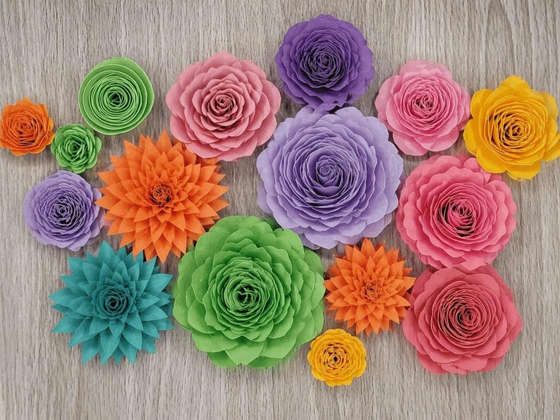 May include: A collection of colorful paper flowers in various sizes and styles. The flowers are in shades of pink, purple, orange, green, yellow, and teal. The flowers are arranged on a light wood-grain surface.
