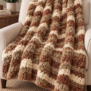 May include: A chunky knit throw blanket featuring a repeating striped pattern in shades of cream, tan, and rust brown. The blanket is crafted from thick, soft yarn with a highly textured, bobble-like appearance, draped over the seat of a chair.