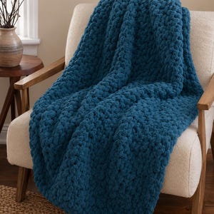 May include: A chunky knit throw blanket in a deep teal blue color. The blanket features a thick, hand-knitted texture with large, soft yarn loops, draped over a cream-colored upholstered chair with wooden armrests and legs.