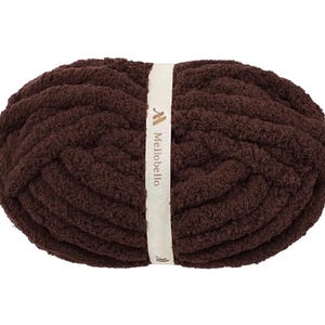 May include: A skein of dark brown, thick, plush yarn wrapped in a cream-colored paper band. The band features the text Mellobello printed in a dark brown font, along with a small logo icon above the brand name and another below it.