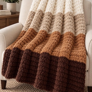 Può includere: Una coperta in maglia grossa caratterizzata da un design a blocchi di colore con strisce orizzontali in crema, beige, marrone chiaro e marrone scuro. La coperta presenta una lavorazione a maglia spessa e testurizzata con un aspetto morbido.