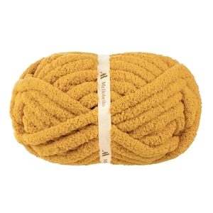 May include: A skein of mustard yellow chunky chenille yarn with a soft, fuzzy texture. A cream-colored paper band wraps around the center of the yarn, featuring the brand name MelloBello printed in dark brown text with a logo icon above it.