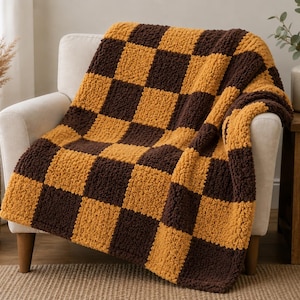 May include: A chunky knit throw blanket featuring a repeating checkerboard pattern in alternating squares of dark brown and mustard yellow. The blanket has a thick, textured yarn construction with a soft, plush appearance draped over a chair.