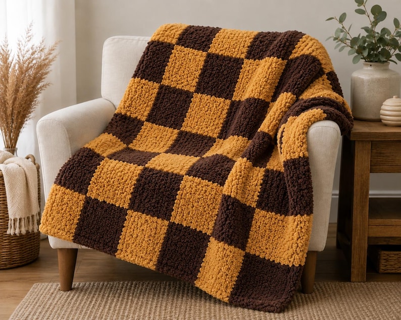 May include: A chunky knit throw blanket featuring a repeating checkerboard pattern in alternating squares of dark brown and mustard yellow. The blanket has a thick, textured yarn construction with a soft, plush appearance draped over a chair.