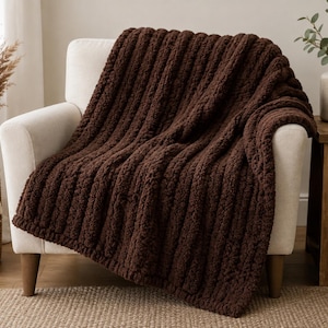 May include: A dark brown chunky knit throw blanket with a prominent vertical ribbed texture. The blanket is draped over a light-colored upholstered armchair, showcasing the thick, soft yarn loops and the consistent, repeating pattern of the knit.