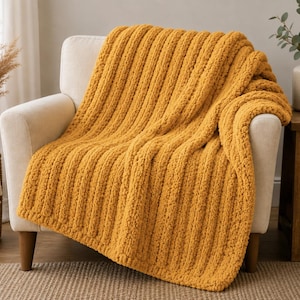 May include: A mustard yellow chunky knit throw blanket with a prominent vertical ribbed texture. The blanket is draped over a light-colored armchair, showcasing its thick, soft yarn construction and consistent, repeating linear pattern throughout.