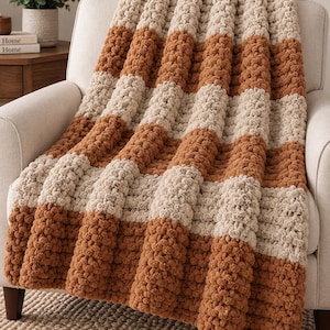 May include: A chunky knit throw blanket featuring thick, soft yarn in alternating horizontal stripes of cream and burnt orange. The blanket has a highly textured, looped appearance and is draped over a neutral-colored upholstered armchair.