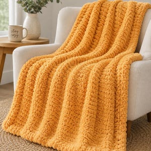 May include: A chunky knit throw blanket in a vibrant mustard yellow colour featuring a thick, ribbed texture. The blanket is draped over a light-coloured armchair, showcasing its soft, plush yarn construction and consistent vertical pattern throughout.