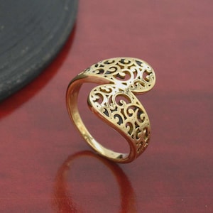 Op de afbeelding: Een goudkleurige ring met een opengewerkt ontwerp. De ring heeft een wervelend, open filigraanpatroon aan de bovenkant, met een band die om de vinger wikkelt. De ring is gemaakt van een glanzend, metalen materiaal.