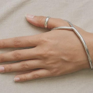 Puede incluir: Una pulsera de mano en tono plateado pulido que presenta una banda de metal lisa y curva que se extiende desde la muñeca hasta un anillo que rodea el pulgar. La pieza tiene un diseño continuo y elegante con un acabado de alto brillo.