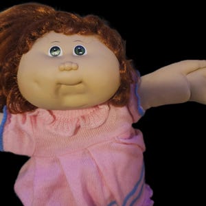 May include: A doll with a round face, dimpled cheeks, and auburn hair styled in a ponytail. The doll has green eyes and is wearing a light pink knit dress with a ruffled collar and blue trim on the sleeves and hemline against a black background.