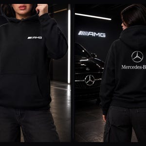May include: Black hoodie with a front pocket and hood. The front features the white AMG logo, and the back displays the Mercedes-Benz logo and text. The hoodie is worn with dark-colored pants.