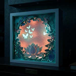 May include: A framed, illuminated paper art piece with a layered design. The artwork features a central lotus flower, butterflies, and foliage in blue, green, and white, set against a warm orange backdrop. The frame is white.