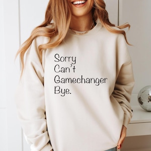 Könnte beinhalten: Cremefarbenes Sweatshirt mit dem Text "Sorry Can't Gamechanger Bye" in Schwarz. Das Sweatshirt hat eine neutrale Farbe und lange Ärmel. Eine dekorative Discokugel ist im Hintergrund zu sehen.