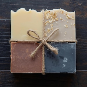 May include: Four bars of handmade soap tied together with twine. The soaps are in shades of cream, brown, and gray, with one bar topped with oats. The soaps are arranged on a dark wooden surface.
