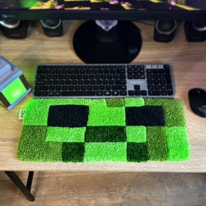 K&ouml;nnte beinhalten: Ein gr&uuml;ner und schwarzer pixeliger Teppich, der an das Gesicht einer Videospiel-Figur erinnert, liegt auf einem Holzschreibtisch. Eine Tastatur und eine Maus sind zu sehen, was auf ein Gaming-Setup hindeutet. Der rechteckige Teppich besteht aus einem strukturierten Material.