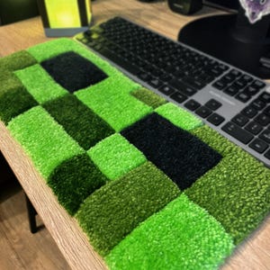 K&ouml;nnte beinhalten: Ein gr&uuml;n-schwarzer, pixeliger Teppich, der an eine Videospiel-Figur erinnert, liegt auf einem Holzschreibtisch neben einer schwarzen Tastatur. Der Teppich besteht aus weichem, strukturiertem Material und hat ein Schachbrettmuster.