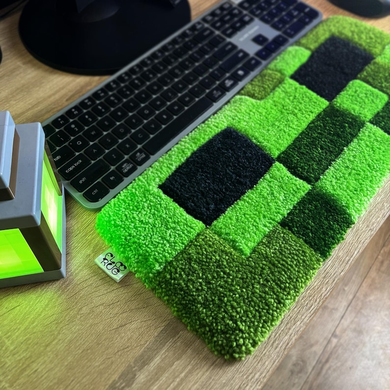 K&ouml;nnte beinhalten: Ein gr&uuml;n-schwarzer, pixeliger Teppich, der einem Videospielcharakter &auml;hnelt, liegt auf einer Holzoberfl&auml;che neben einer Tastatur. Der Teppich hat ein strukturiertes, getuftetes Aussehen. Ein kleines Etikett mit dem Text "CLUB RUG" ist am Teppich befestigt.