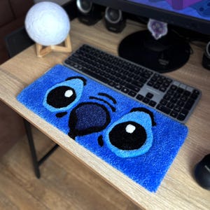 Op de afbeelding: Een blauw rechthoekig tapijt met het gezicht van een stripfiguur, met grote zwarte ogen en een donkerblauwe neus. Het tapijt ligt op een houten bureau, naast een toetsenbord en een witte bolvormige decoratie.