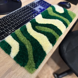 Könnte beinhalten: Eine rechteckige, getuftete Schreibtischunterlage mit einem wellenförmigen Muster in Grün- und Cremetönen. Die Unterlage liegt auf einem hellen Holzschreibtisch neben einer Tastatur und einer Maus. Das Design ähnelt einer stilisierten Landschaft.