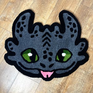 Peut inclure: Un tapis gris en forme de tête de dragon, avec des yeux verts et une langue rose. Le tapis a un contour noir et des détails noirs. Il est posé sur un sol en bois.