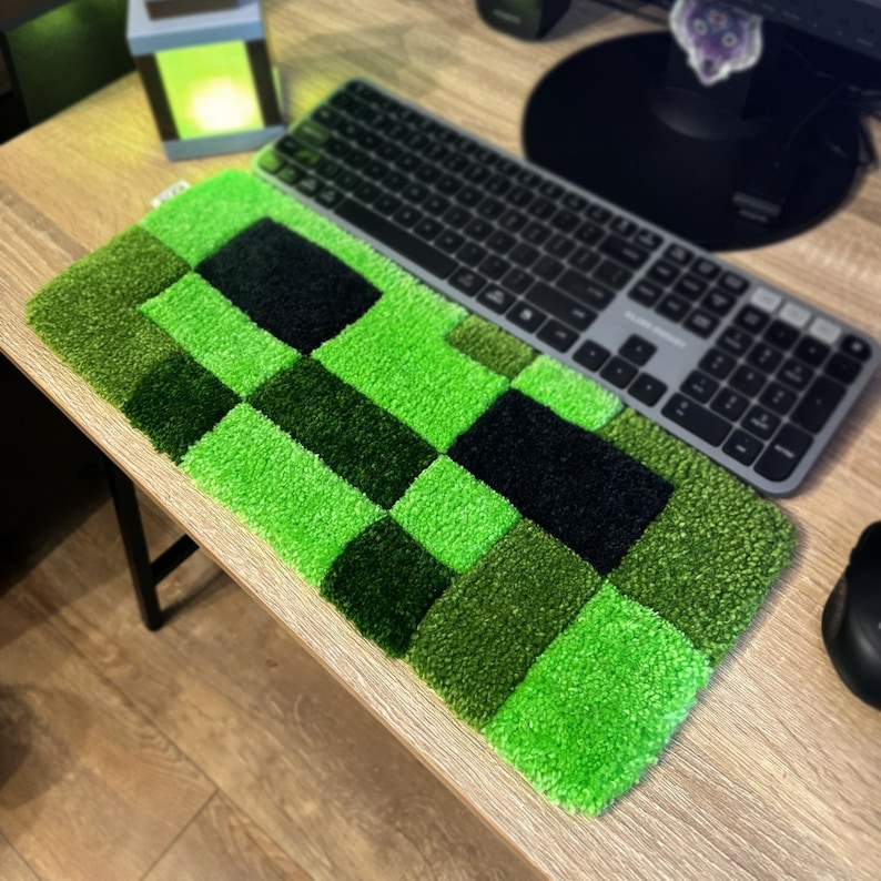 K&ouml;nnte beinhalten: Ein rechteckiger, pixeliger Teppich in Gr&uuml;n- und Schwarzt&ouml;nen, der an eine Spielfigur erinnert. Eine graue Tastatur liegt auf dem Teppich. Der Teppich liegt auf einem Holztisch, im Hintergrund eine kleine Lampe.