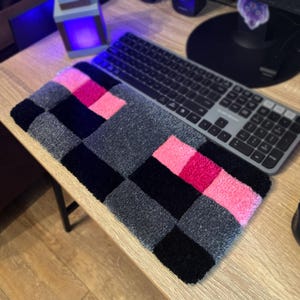 Könnte beinhalten: Handgefertigte Tastaturmatte mit Schachbrettmuster. Das Design zeigt Quadrate in Schwarz, Grau und Rosa. Eine graue Tastatur liegt auf der Matte, die auf einem Holztisch platziert ist.