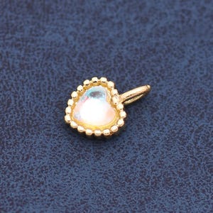 May include: A small heart-shaped charm featuring a central iridescent stone set within a gold-toned metal frame. The edge of the heart is decorated with a delicate beaded texture, and a small gold-toned jump ring is attached to the top side.